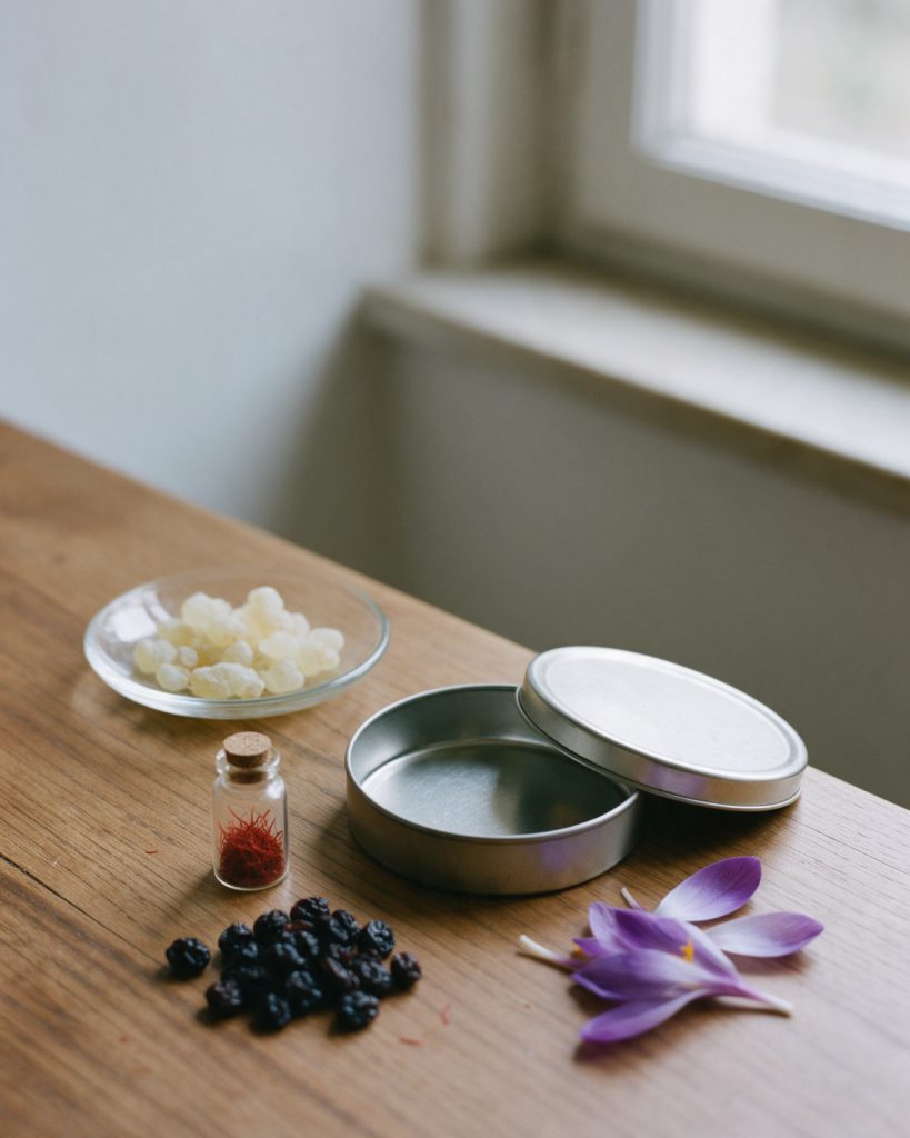 An open plain round tin beside a glass dish of mastic tears, a small vial of red saffron, a heap of currants and violet crocus petals on a wooden table